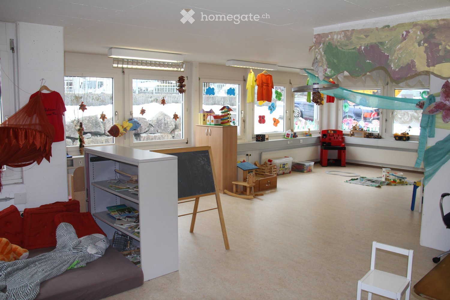 A brightly lit playroom with a variety of toys and furniture, including a sofa, shelves, a rocking horse, and a blackboard. Large windows provide natural light, and the space is decorated with colorful artwork and hanging decorations.