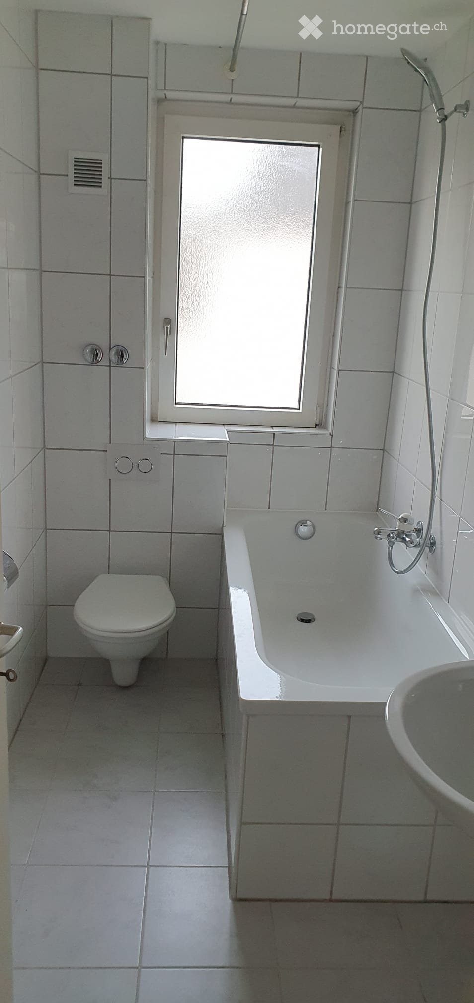 Modern bathroom with white tiles, a bathtub, toilet, and sink. Features a window above the bathtub and a small cabinet with mirrored doors.