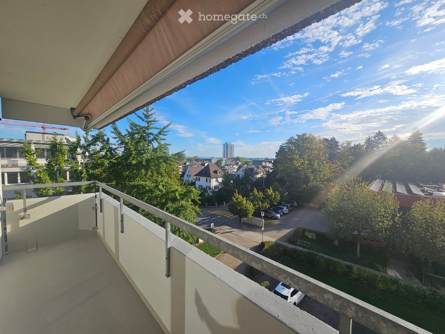 Balcony with metal railing, overlooking a residential area with trees, parked cars, and distant buildings under a partly cloudy sky.