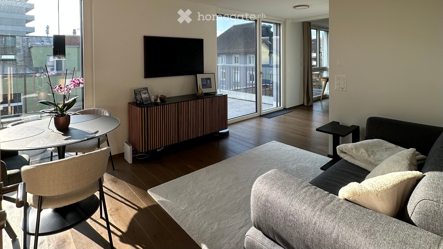 Modern living room with a grey sofa, coffee table, dining table, chairs, TV, and a balcony view.