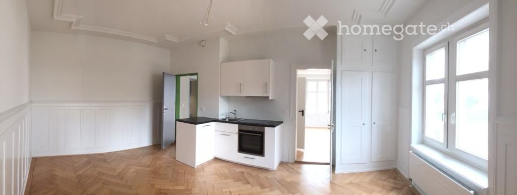 Bright, unfurnished room with white walls, wooden flooring, and built-in white cabinets. Features a small kitchen area with a sink, oven, and countertop.