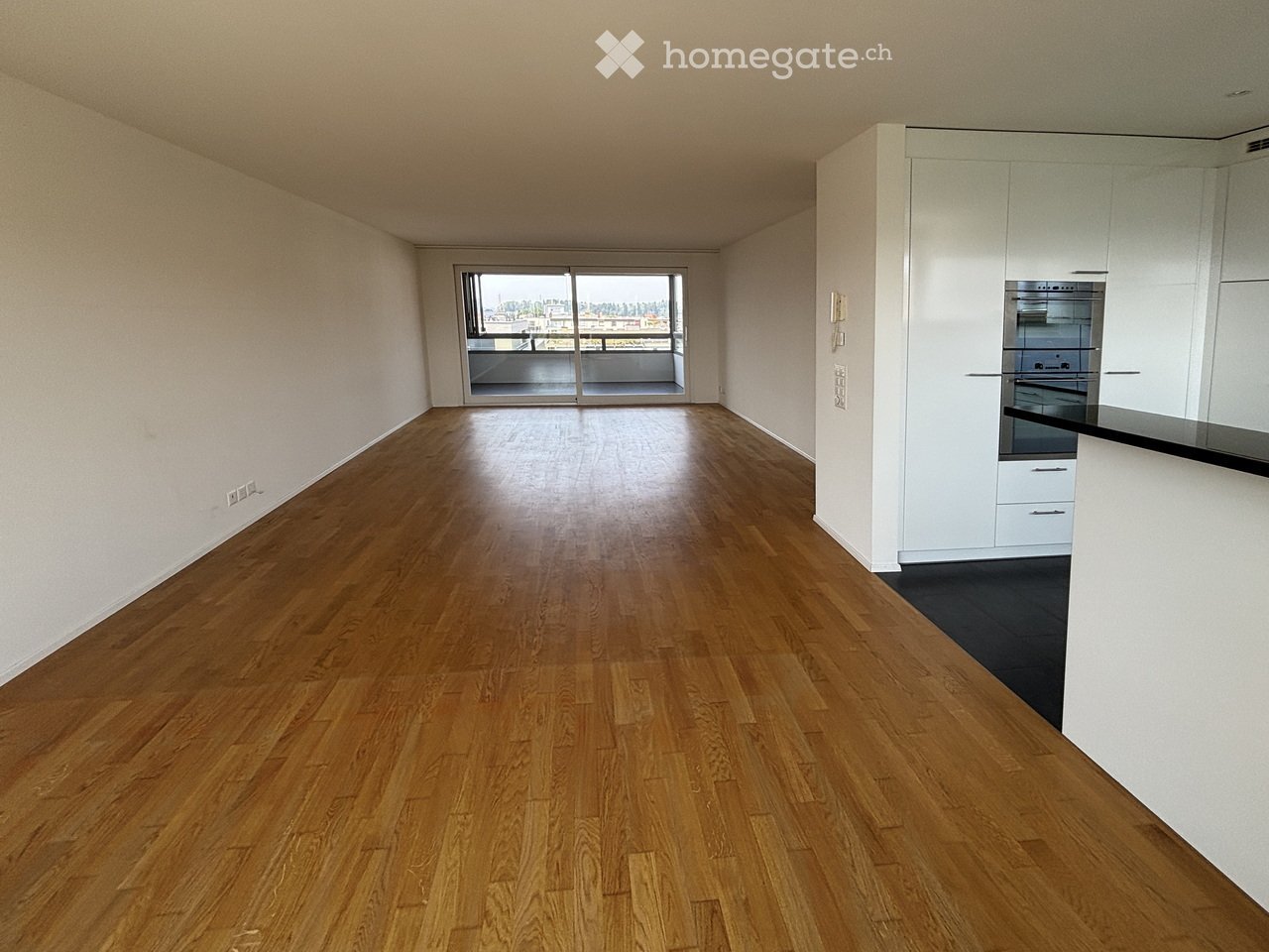 Empty room with wooden flooring, white walls, and a sliding glass door leading to a balcony. A small kitchen area with white cabinets and stainless steel appliances is visible on the right side.