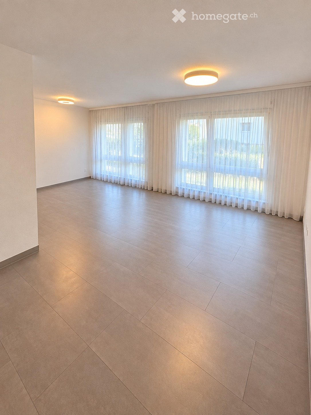 Empty room with tiled floor, white walls, large windows with sheer curtains, and recessed lighting.