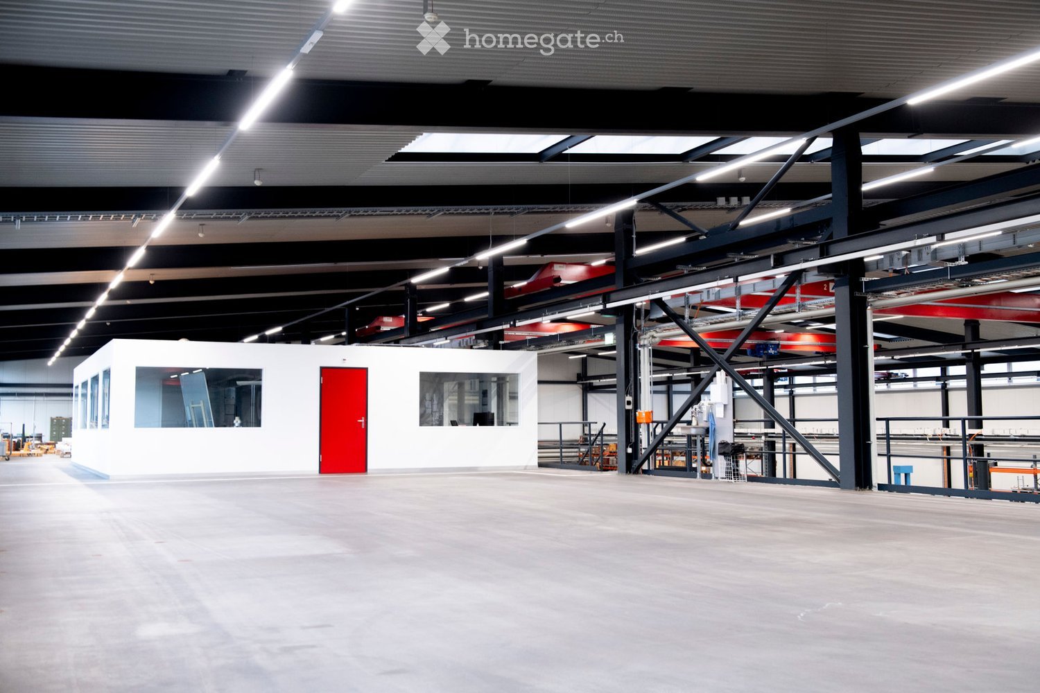 Large industrial-style building with a spacious, open floor plan, high ceilings, exposed metal beams, and bright lighting. Features a small white room with a red door and windows, along with various industrial equipment and storage areas.