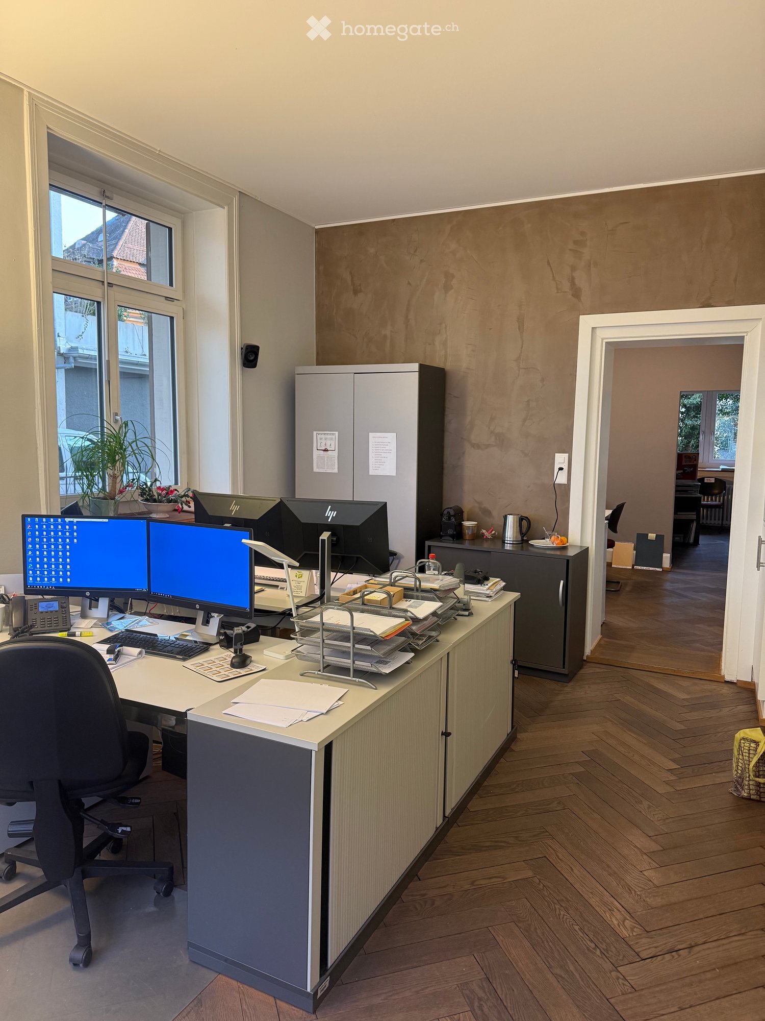 Office room with three computer screens, black office chair, gray desks, beige cabinets, radiator on the wall, and plants on the window sill