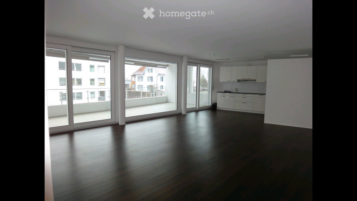 Empty room with dark wooden flooring, large glass sliding doors leading to a balcony, and a small kitchen area with white cabinets.