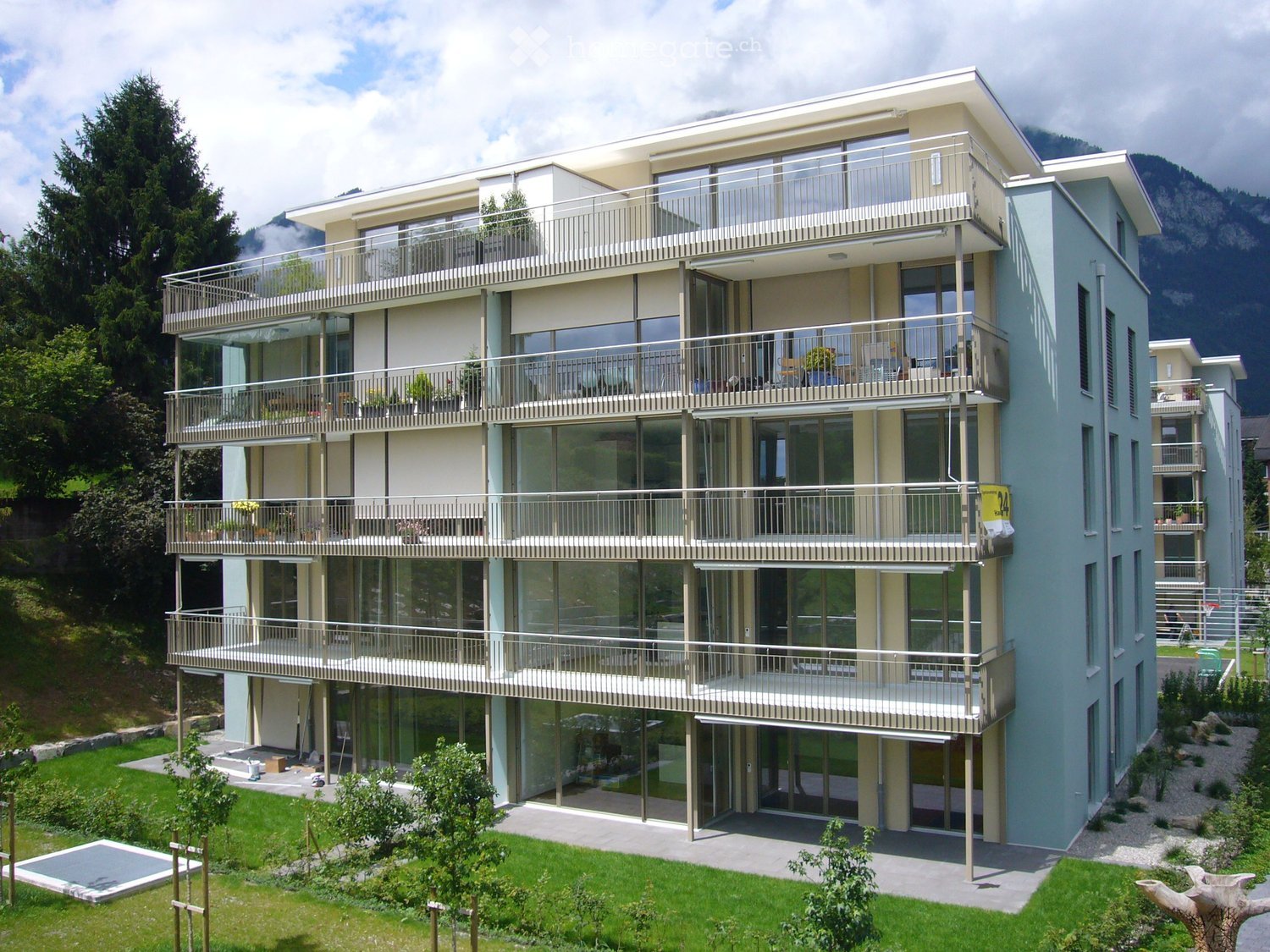 Modern multi-story building with large glass windows and balconies, surrounded by greenery and a swimming pool.