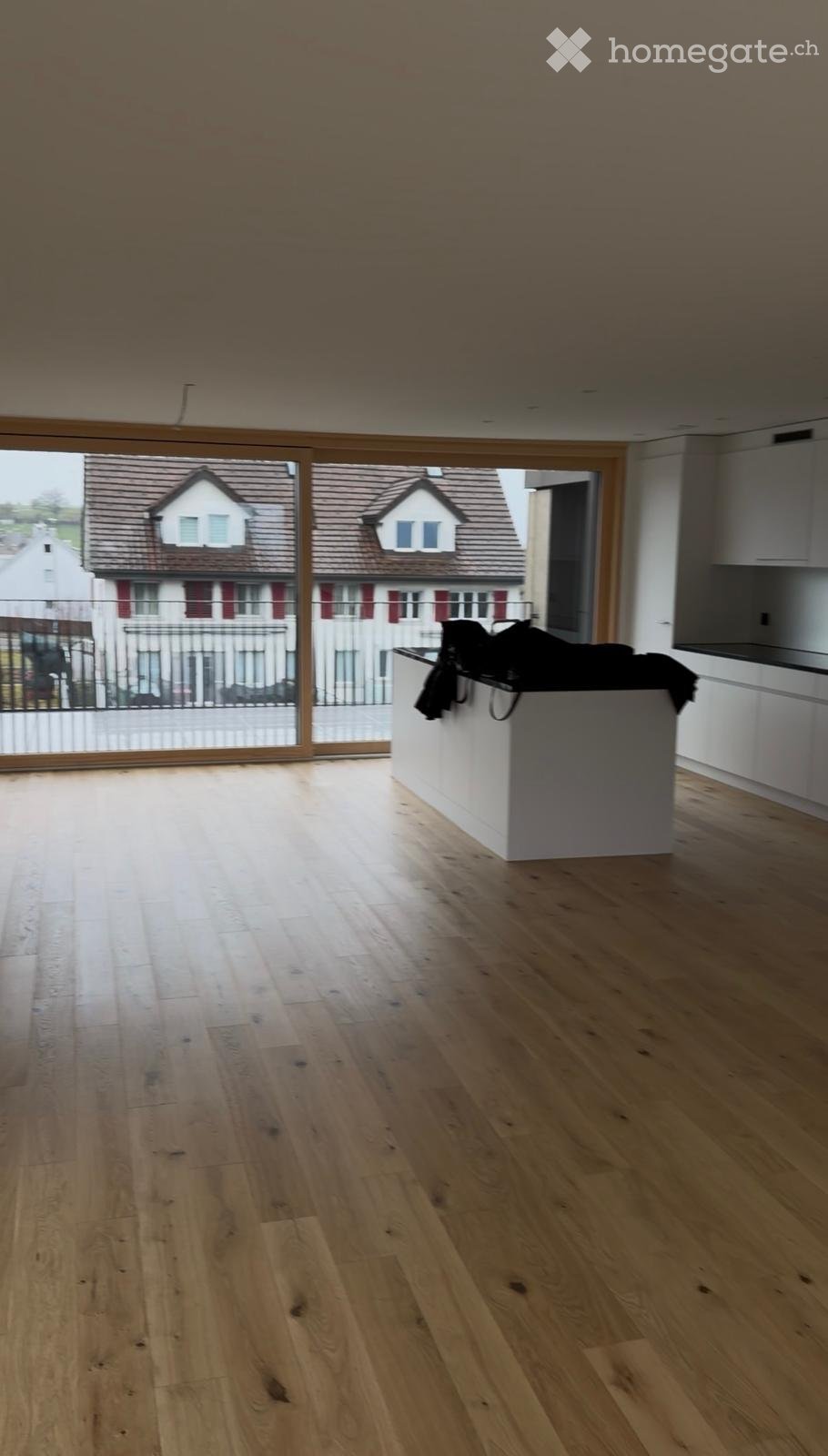Empty room with light wooden flooring, white kitchen counter, and large windows overlooking a residential area.