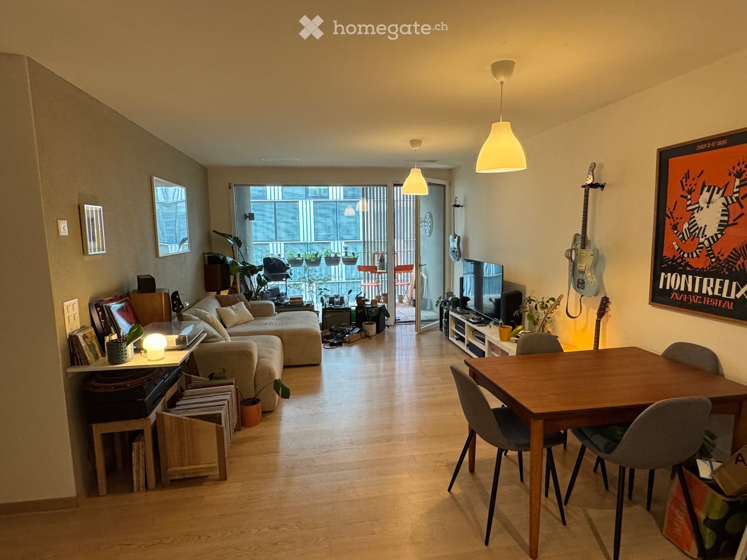 A modern, open-concept living and dining area with a balcony. Features include a grey sofa, ottoman, wooden dining table with chairs, a record player, guitars on the wall, and a television. The space is well-lit with pendant lights and has a view of the b
