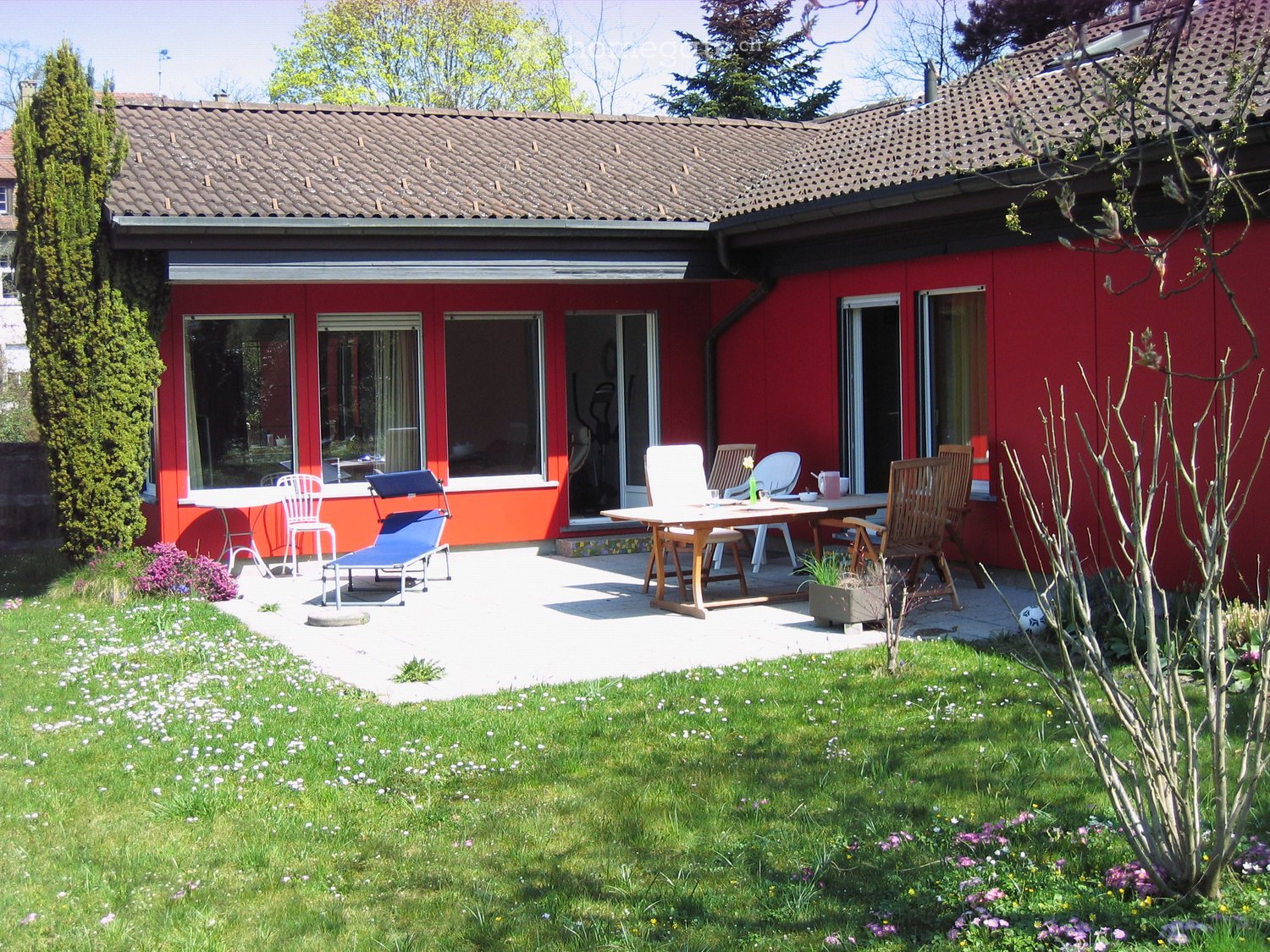 Garden with a patio area featuring a table, chairs, and a lounge chair, surrounded by grass and flowers.