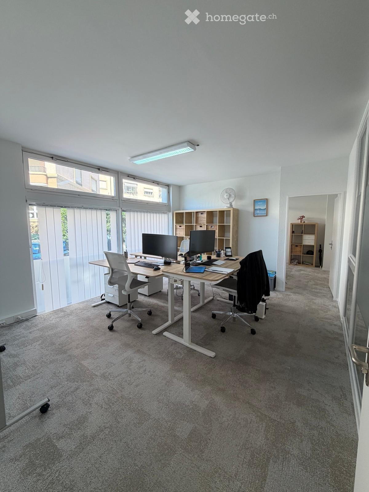 Bright, modern office space with large windows, a standing desk, two chairs, dual monitors, and shelves for storage.