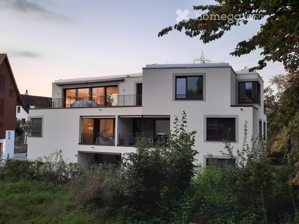 Modern two-story house with white exterior, large windows, and balconies. The ground floor features a garage entrance, and the upper level has expansive windows and a balcony.