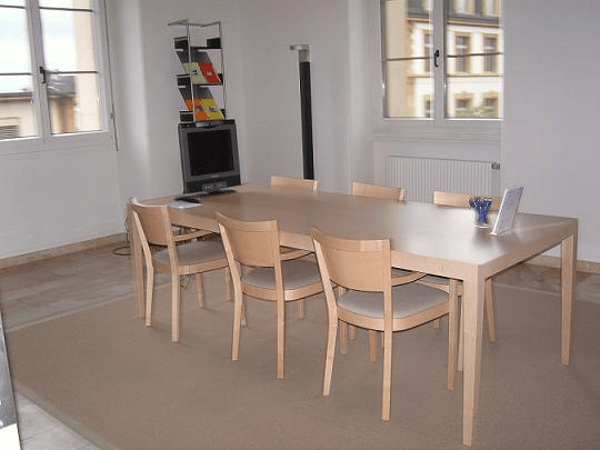 A dining room with a long wooden table, chairs, a monitor, a mouse, a book, and a pen holder on the table. The room has white walls, a radiator, and large windows with a view of the outside.