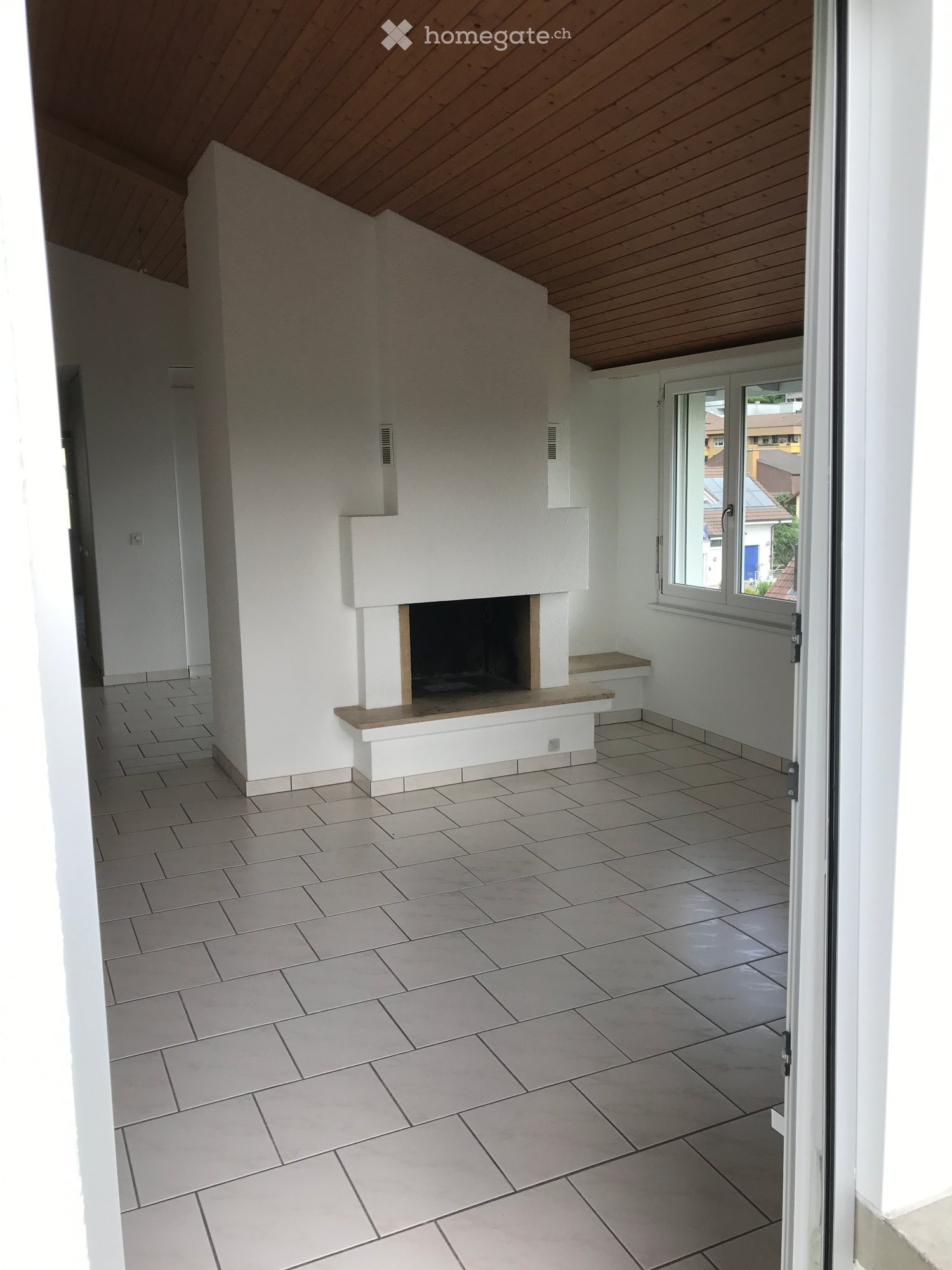 Empty room with white walls, tiled floor, and a built-in fireplace with a wooden bench. The room has a slanted wooden ceiling and a large window with a view of the outside.