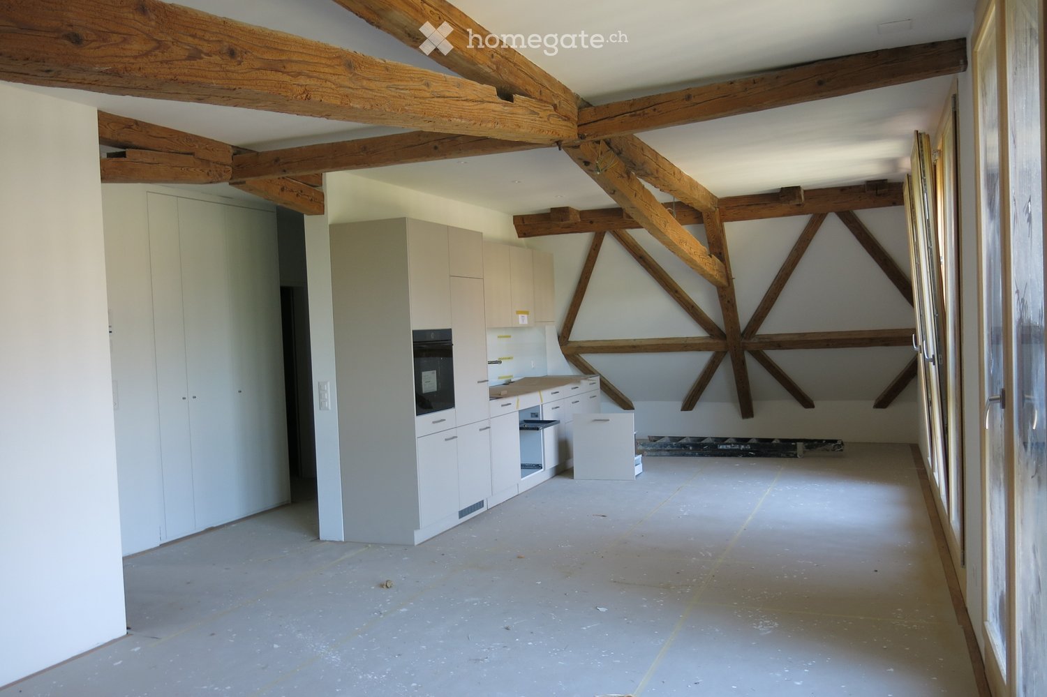 Empty room with exposed wooden beams, white cabinetry, and a partially constructed kitchen area. The floor is unfinished concrete, and the walls are painted white.