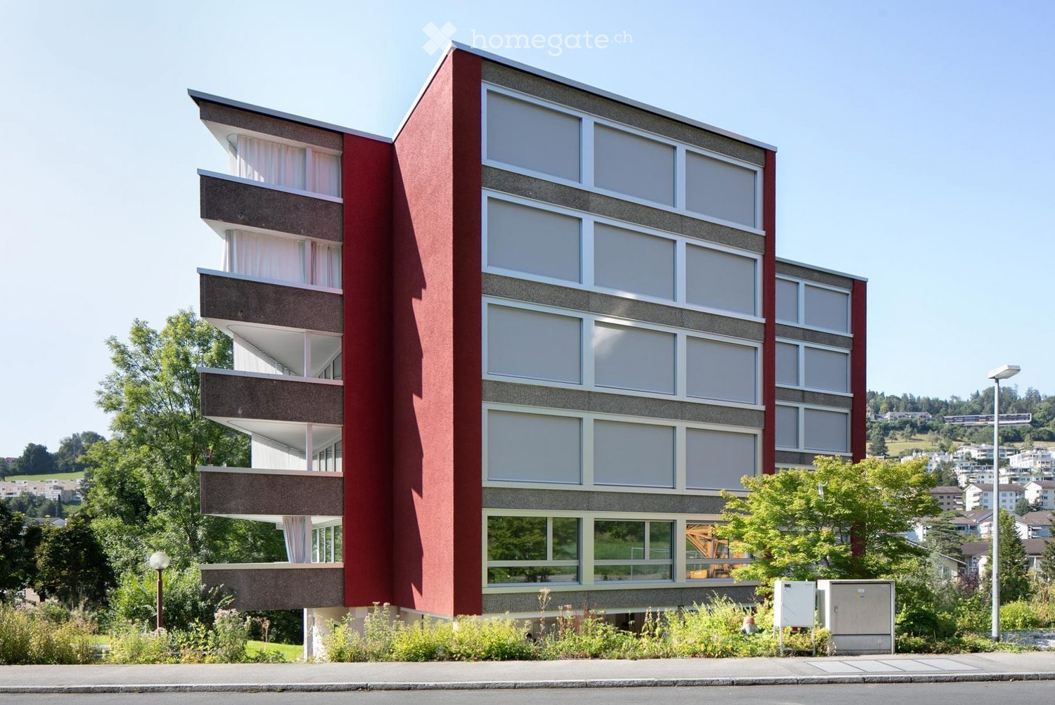Modern multi-story building with red and gray facade, large windows, and balconies. Surrounded by greenery and trees.