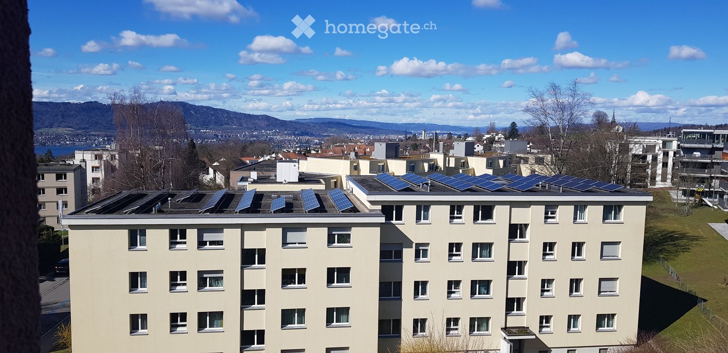 Multi-story residential building with solar panels on the roof, surrounded by other buildings and a scenic view of mountains and a lake in the background.