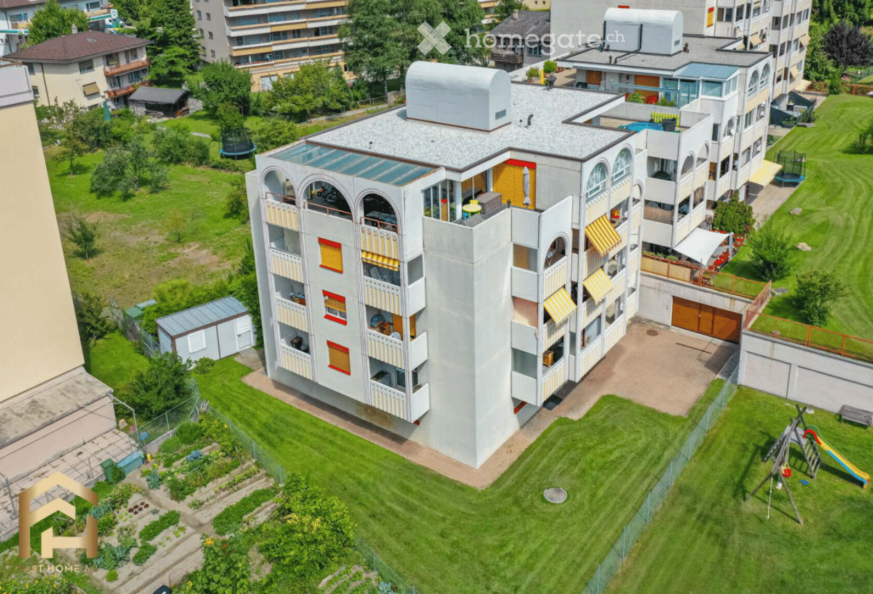 Aerial view of a modern multi-story residential building with balconies, surrounded by green lawns, trees, and playground equipment.