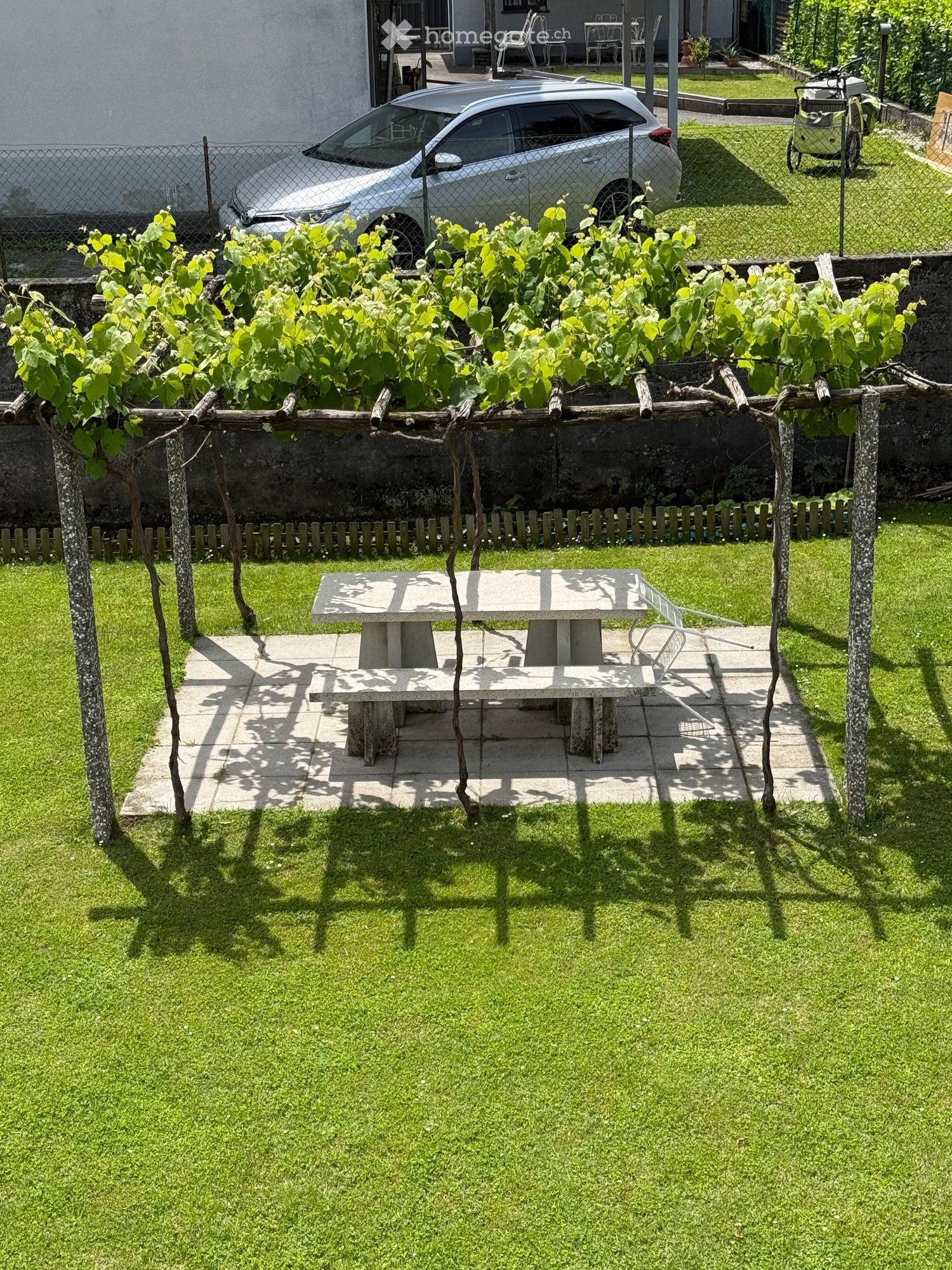 Well-maintained garden with a stone table and chair, surrounded by green grass and a wooden fence, with a grapevine trellis and a parked car in the background.