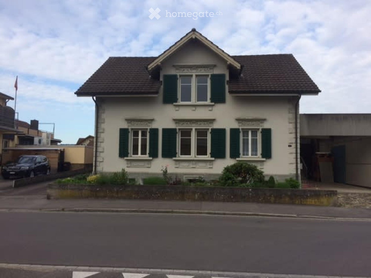 Two-story house with beige exterior, green shutters, and a small garden in front.