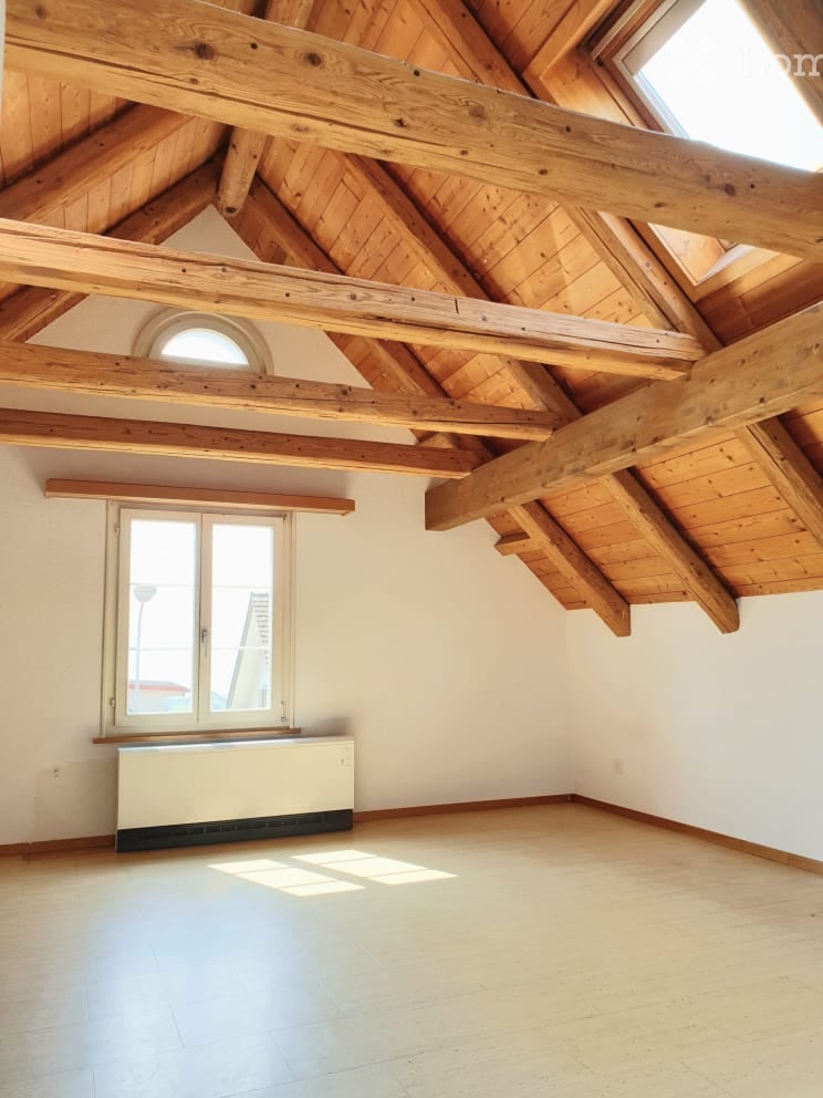 Empty attic space with exposed wooden beams, skylights, and a large window. The room has a polished wooden floor and white walls.