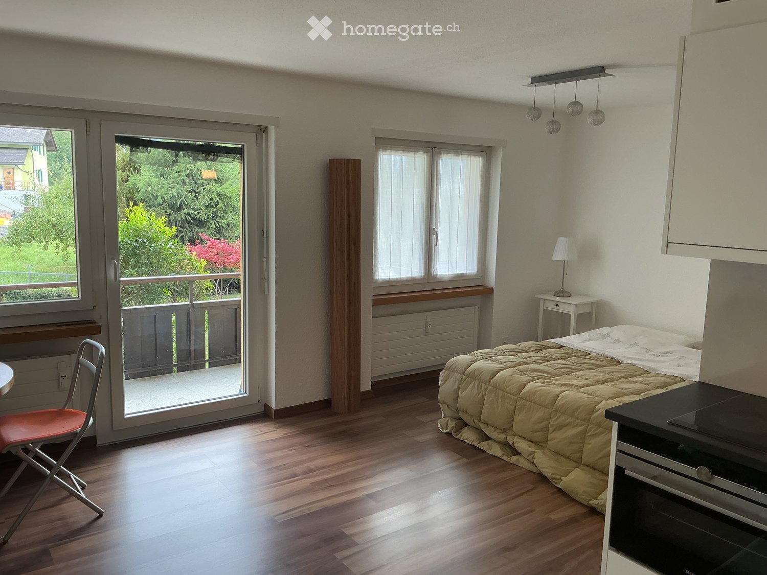 Bright bedroom with wooden flooring, large sliding glass door leading to a balcony, bed with beige duvet, small table with lamp, and a red folding chair.