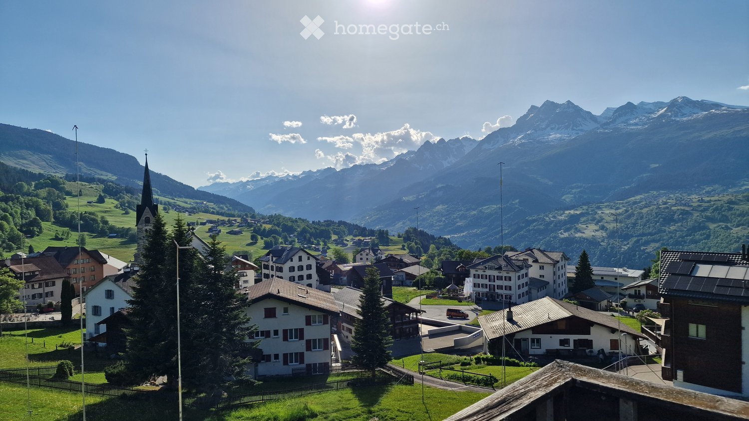 A picturesque village nestled in a valley with traditional houses, surrounded by lush green fields and towering mountains under a clear blue sky.