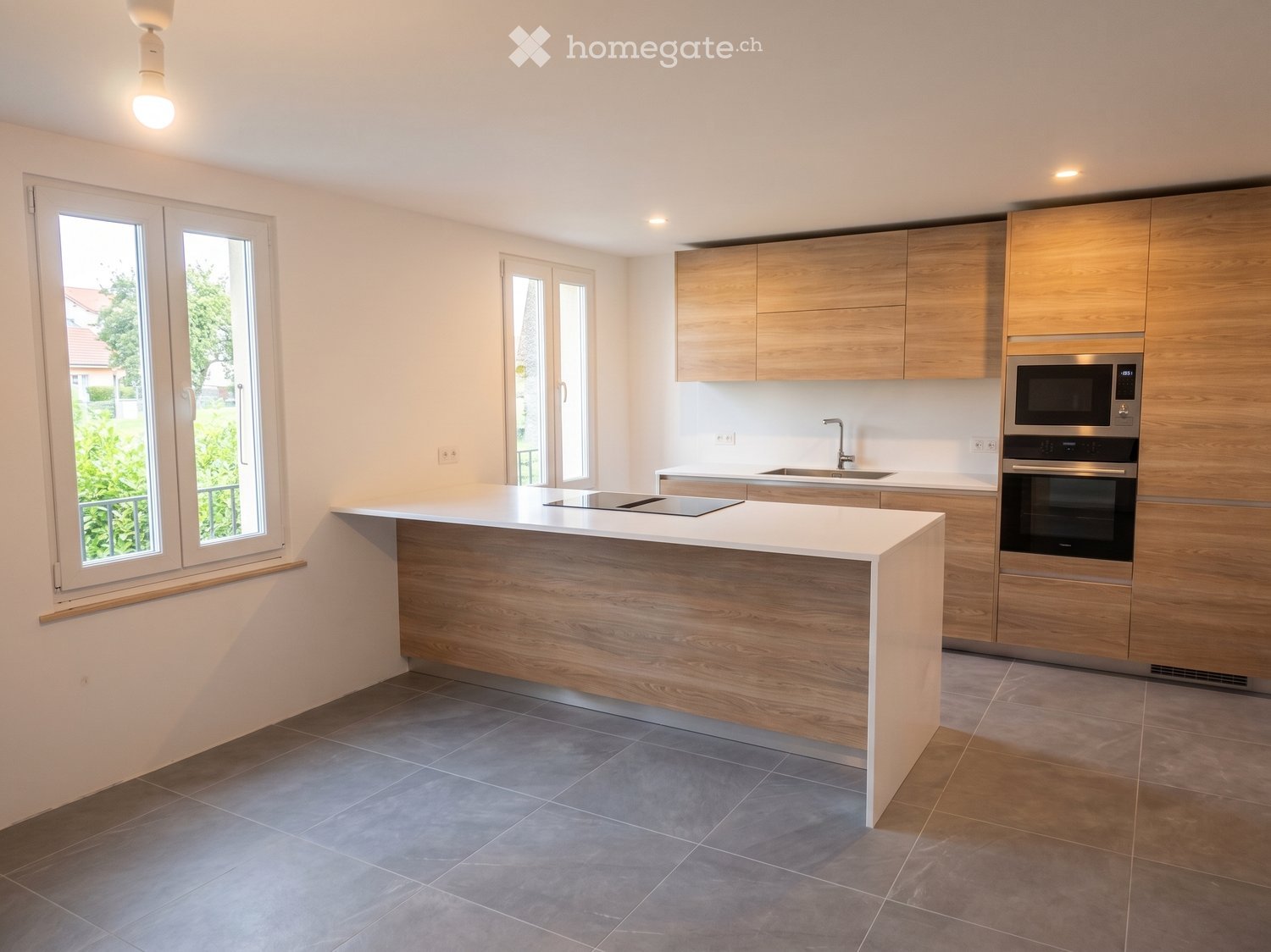 Modern kitchen with light wooden cabinets, white countertops, and stainless steel appliances including a microwave and oven. Features a central island with a built-in induction cooktop and ample natural light from large windows.