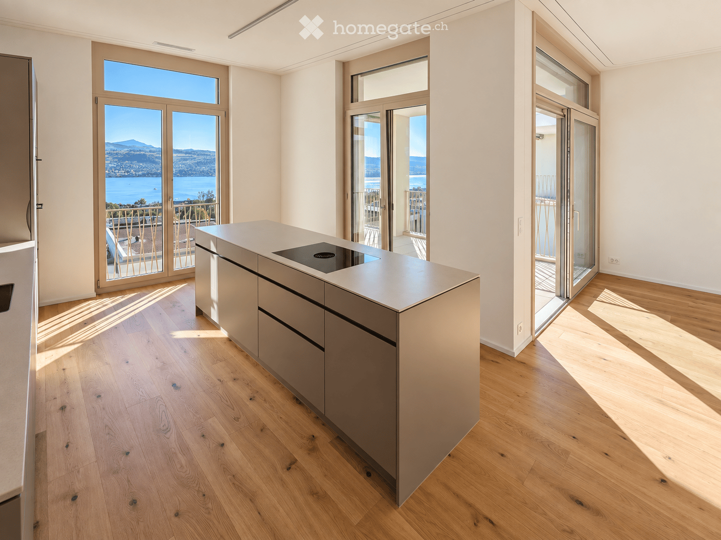 Modern kitchen with light wooden flooring, a central island with an induction cooktop, and large glass doors opening to a balcony with a scenic view of a lake and mountains.