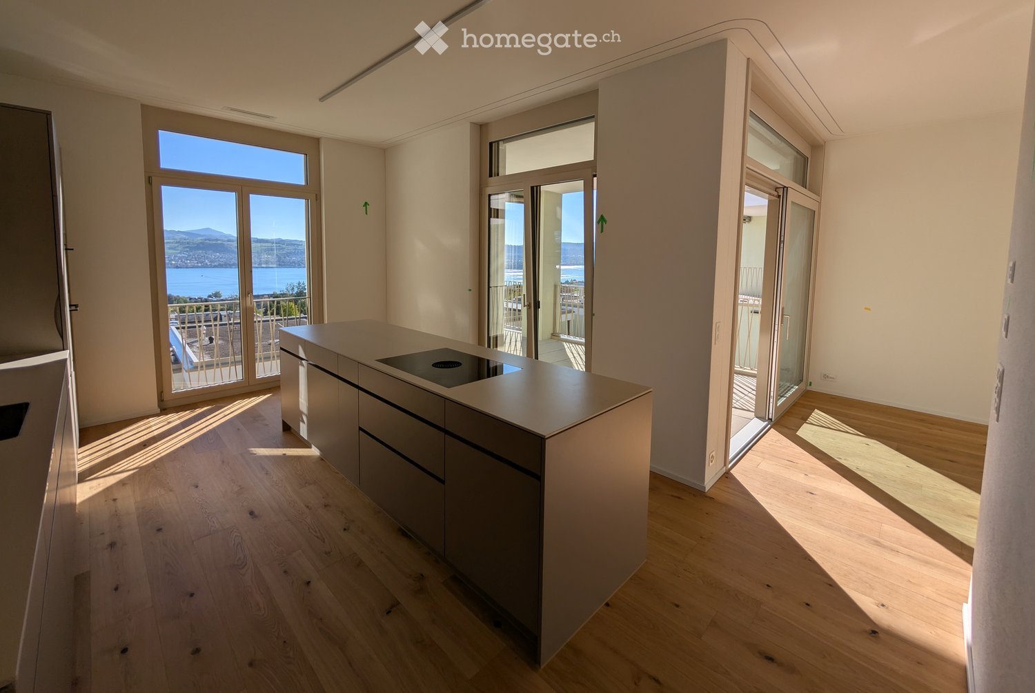 Modern kitchen with a central island, light wood flooring, and large windows offering a scenic view of the water and mountains.