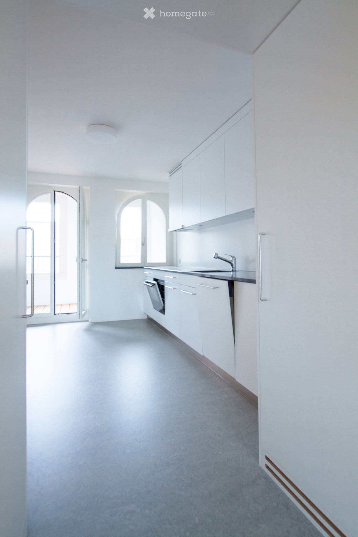 Empty kitchen with white cabinets, stainless steel sink, and dishwasher. Large arched windows and doors provide natural light.
