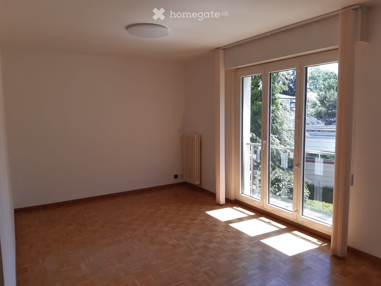 Empty room with wooden flooring, large sliding glass doors leading to a balcony, and a radiator on the wall.