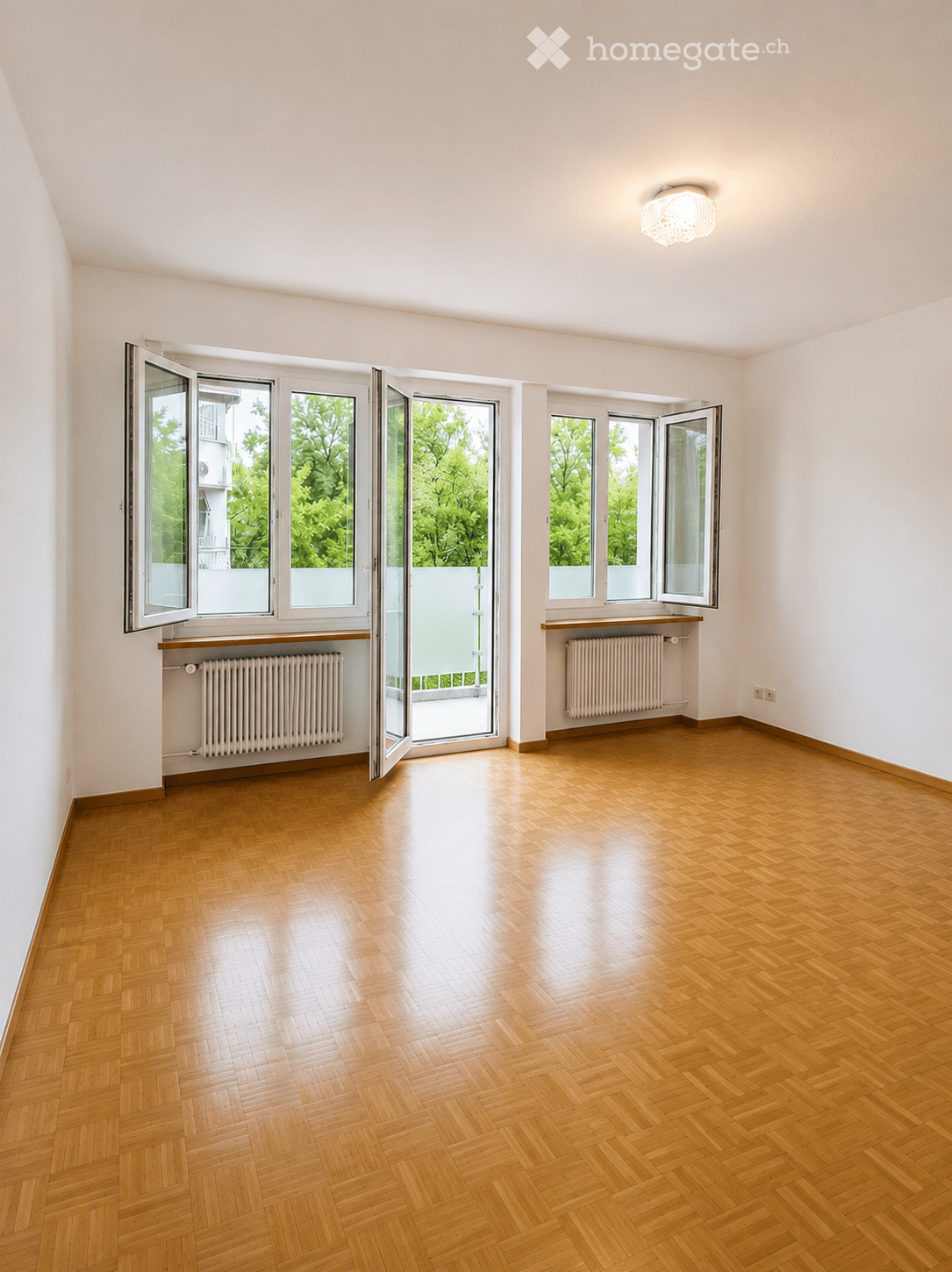 Empty room with wooden flooring, large windows, radiators, and a ceiling light fixture.