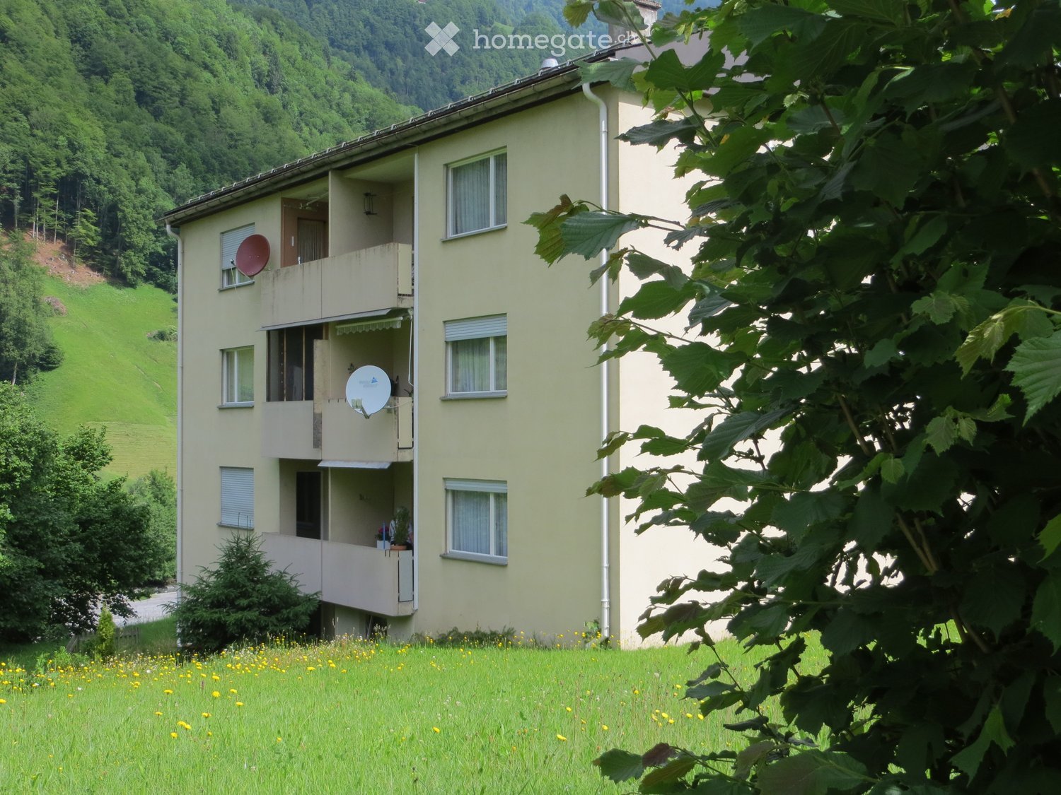 A three-story residential building with balconies, satellite dishes, and a view of a green, hilly landscape.