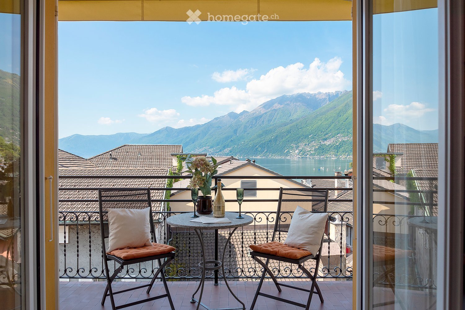 Balcony with a stunning view of the mountains and lake, furnished with a small table and two chairs, decorated with flowers and wine bottles
