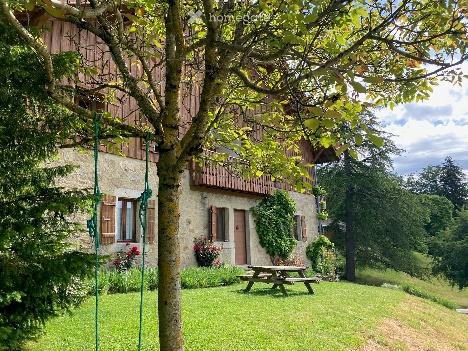 A rustic stone house with wooden accents, a balcony, and a well-maintained garden with a picnic table and swing.
