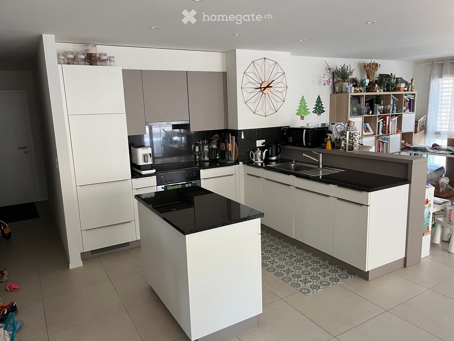 Modern kitchen with white cabinets, black countertops, stainless steel appliances, and a central island. Features a sink, microwave, coffee maker, and decorative items on shelves and countertops.