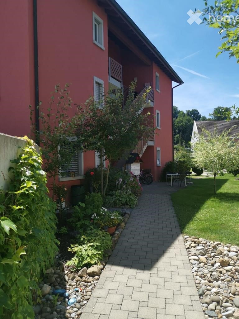 Red brick two-story building with white-framed windows, a staircase leading to a balcony, and a well-maintained garden with a brick pathway, small table, and chairs.
