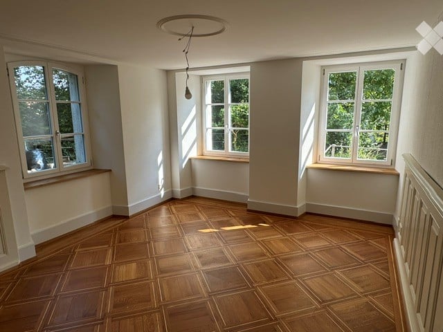 Empty room with wooden flooring, white walls, and three large windows allowing natural light to enter. The room features a simple, clean design with no furniture or decorations.