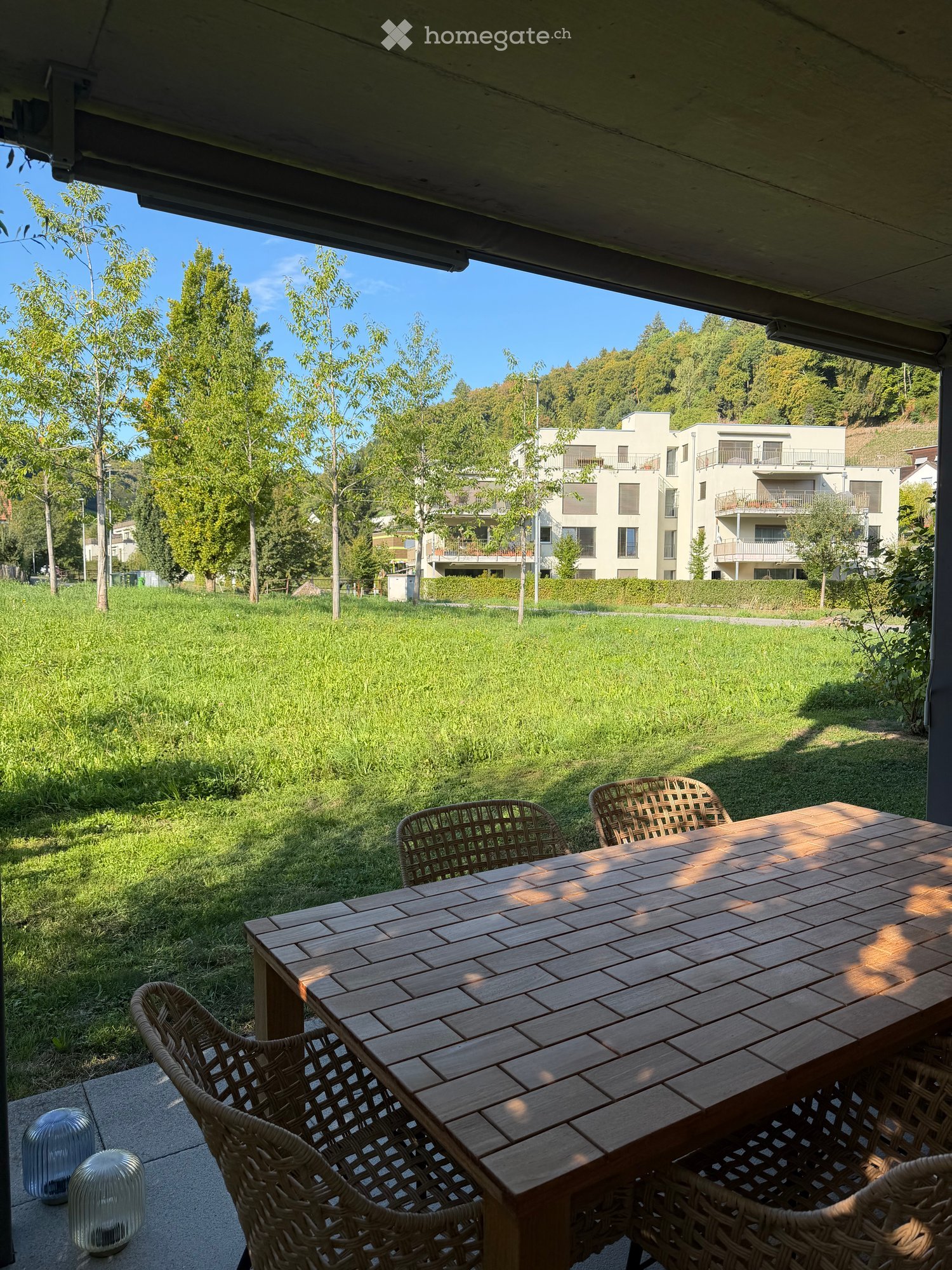 Balcony with wooden table and wicker chairs, overlooking a grassy area with trees and modern apartment buildings.