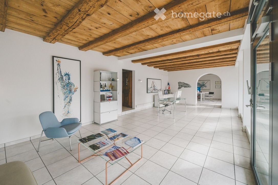 Bright, open living space with white tiled flooring, wooden ceiling beams, and large windows. Features a light blue armchair, glass coffee table with books, white shelving unit, and a workspace area with a desk and laptop.
