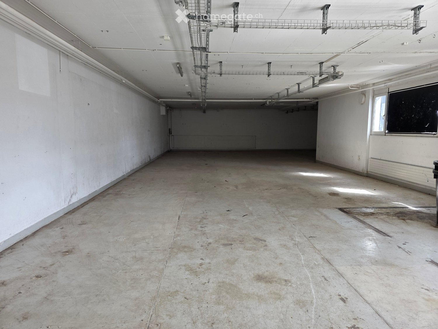 Empty room with bare concrete floor and white walls, exposed ceiling with metal shelving units, and a small window on the right side.