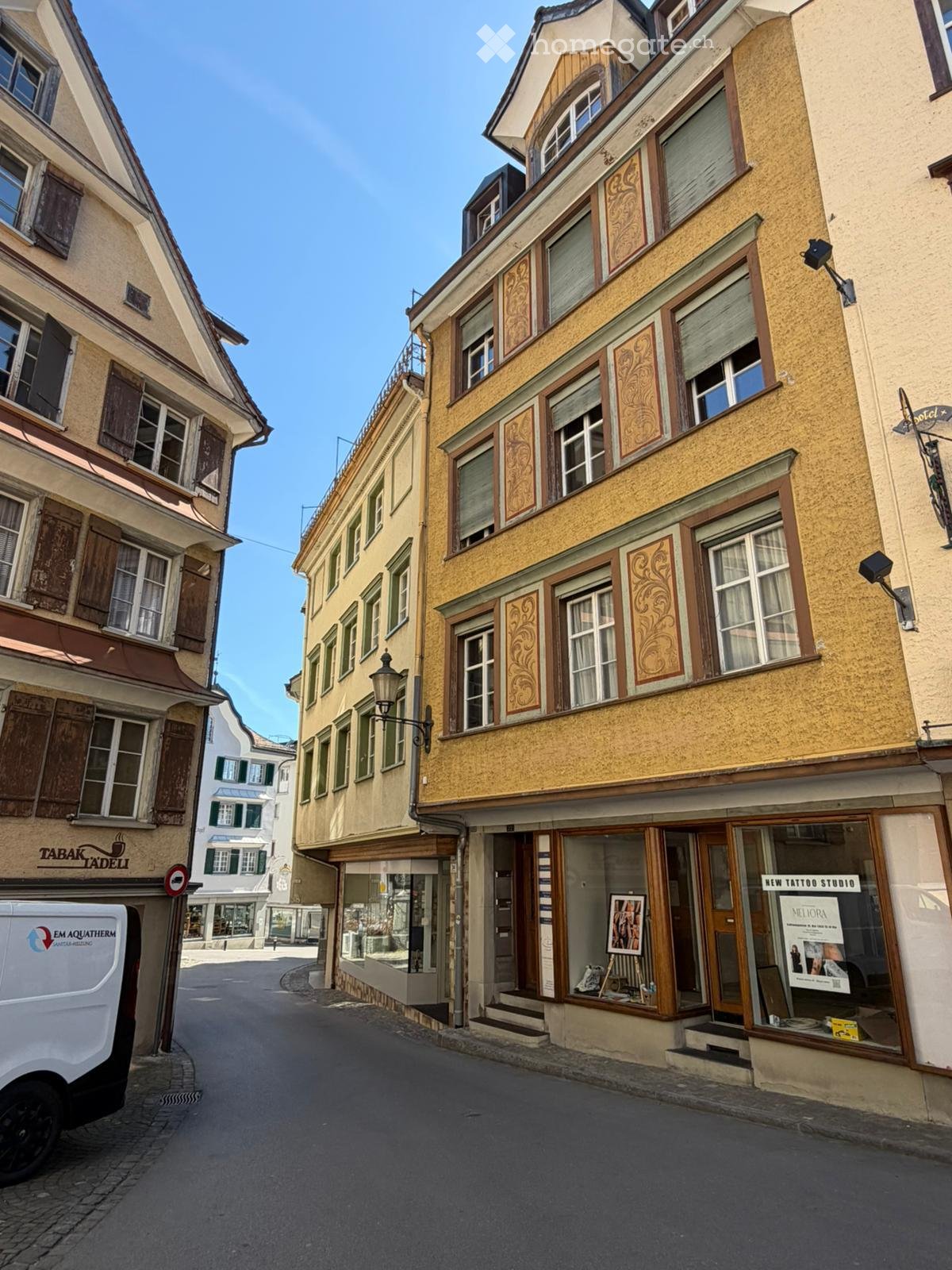 A narrow street lined with multi-story buildings featuring ornate wooden facades, large windows, and shutters. Shops with glass storefronts are visible, including a tattoo studio and a coffee shop.