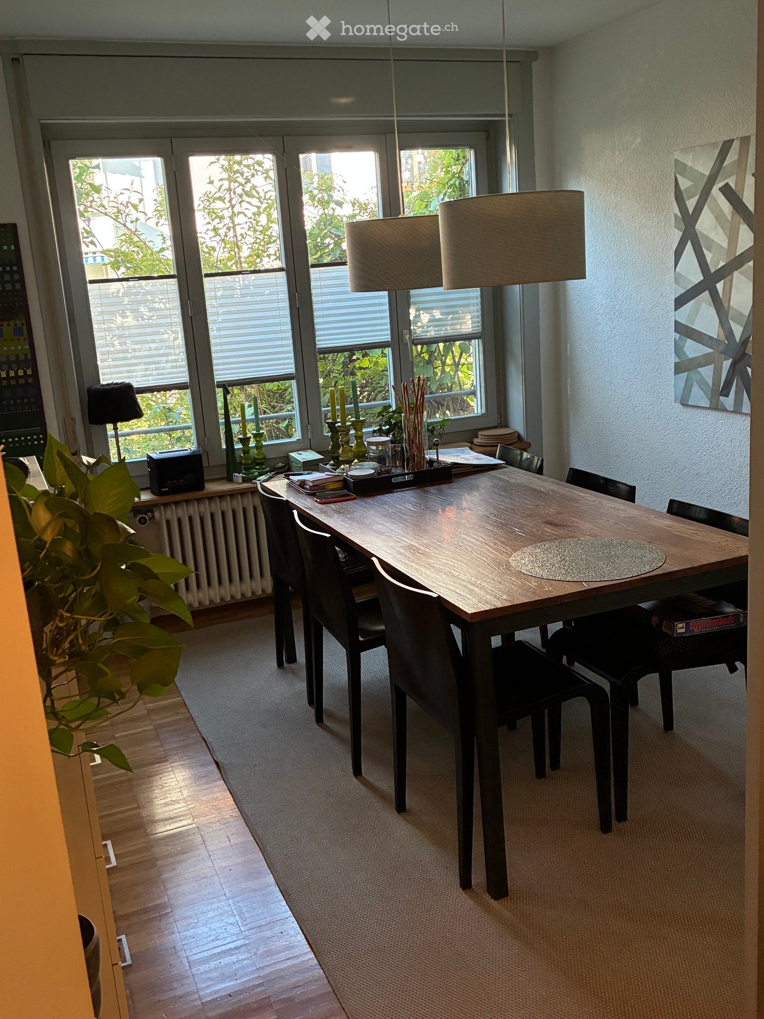 Bright dining area with a wooden dining table, black chairs, hanging lamps, and decorative candles on a side table. Large windows with blinds offer natural light and a view of greenery.