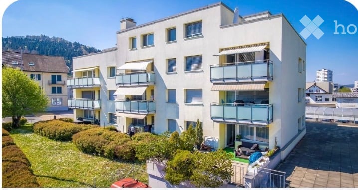 Modern multi-story apartment building with multiple balconies and awnings, surrounded by greenery and a well-maintained lawn.
