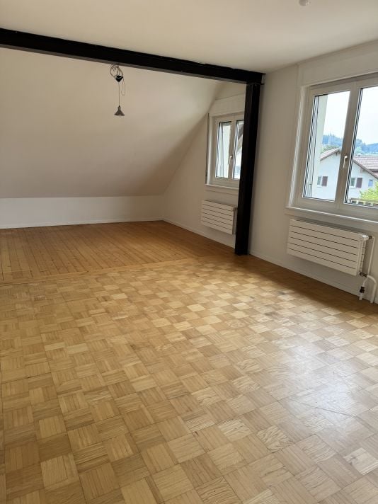 Empty room with wooden parquet flooring, white walls, and large windows allowing natural light. The room features a slanted ceiling typical of an attic space.