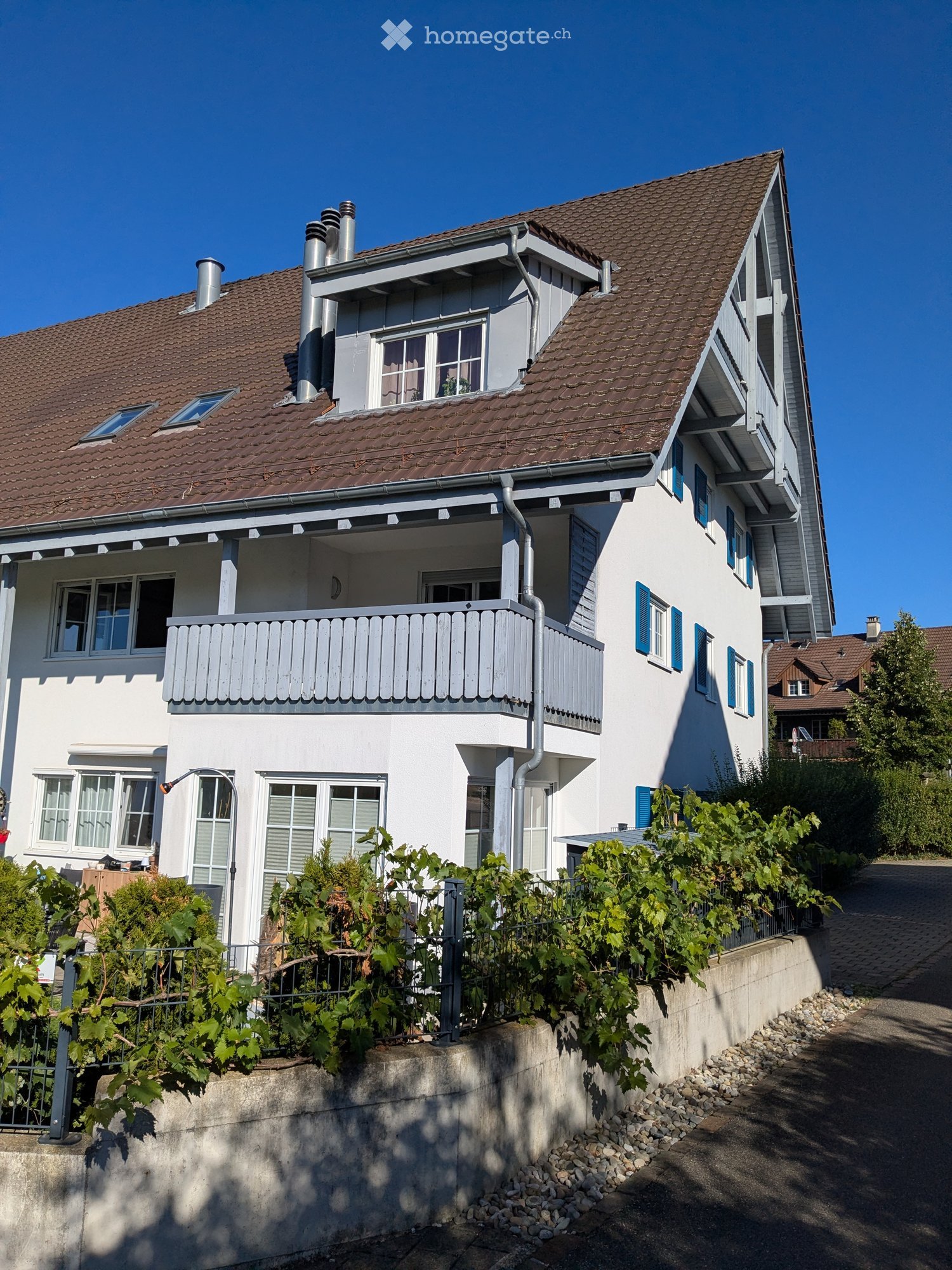 Two-story house with a red-tiled roof, white walls, blue shutters, and a balcony on the first floor. The house has multiple windows and is surrounded by greenery and a low concrete wall.
