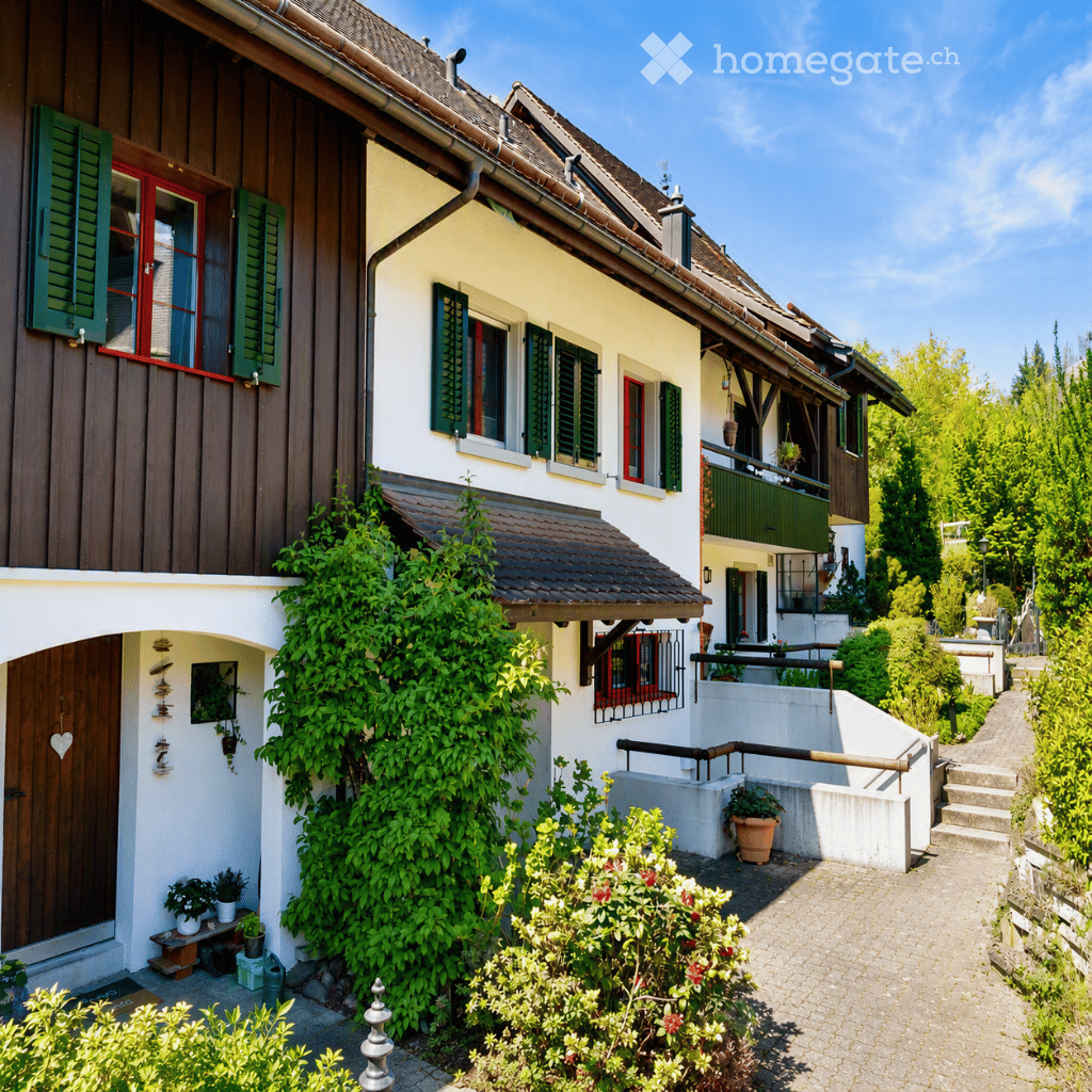 Two-story house with white walls, red-framed windows, green shutters, and a tiled roof. The house features a wooden door, climbing plants, and a small garden area with potted plants and a pathway.