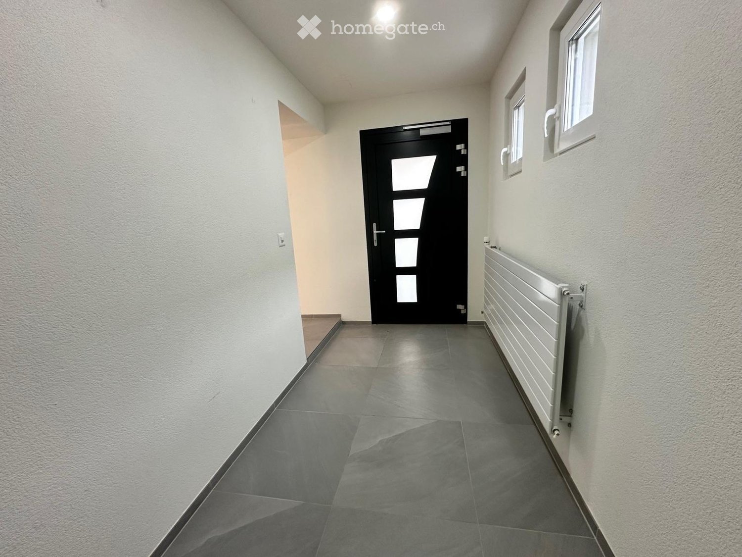 Empty hallway with grey tiled floor, white walls, a black door with glass panels, and a radiator on the right side.