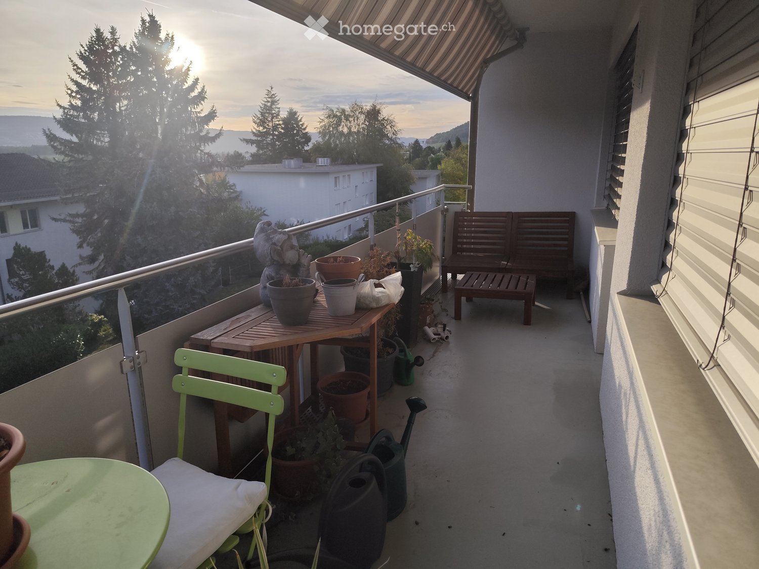 Balcony with a wooden table, green chair, wooden bench, potted plants, and garden hoses.